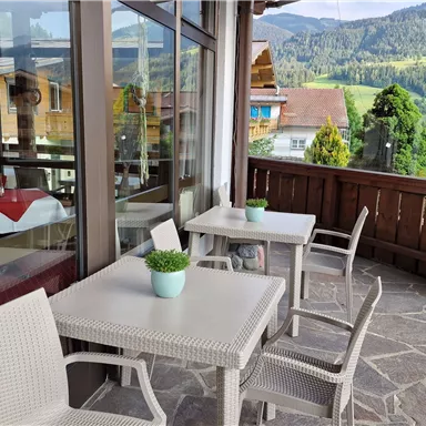 A cozy outdoor area with white tables and chairs. In the background, green mountains and a tranquil landscape are visible.