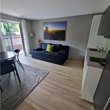 Ein modernes Wohnzimmer mit einem schwarzen Sofa und einem runden Tisch. Eine große Fensterfront bringt viel Licht und bietet einen Zugang zu einem Balkon.