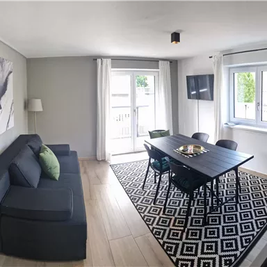 Ein modernes Wohnzimmer mit einem dunklen Sofa und einem Esstisch. Große Fenster sorgen für viel Tageslicht und einen gemütlichen Aufenthalt.