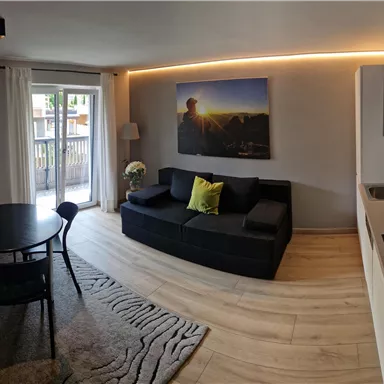 Ein modernes Wohnzimmer mit einem schwarzen Sofa, einem Essbereich und einer offenen Küche. Große Fenster lassen viel Licht herein und ein Kunstwerk hängt an der Wand.