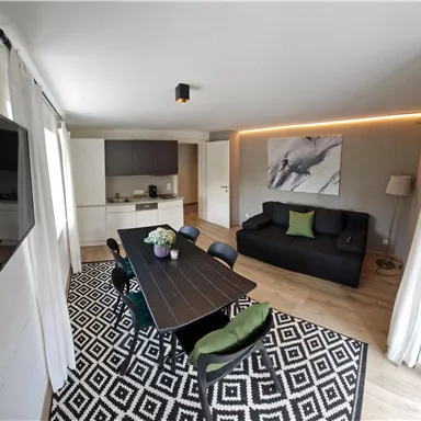 Ein modernes Wohn-Esszimmer mit einem großen Tisch, schwarzen Stühlen und einer gemütlichen Couch. Die Wände sind hell, und es gibt eine elegante Teppichgestaltung.