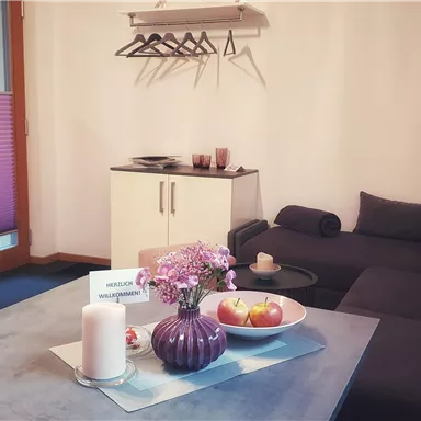A cozy living space with a sofa, a table, and a modern kitchen corner. Fresh fruits and a flower vase are on the table.