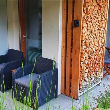 A cozy outdoor area with two black armchairs and a small table. The walls are adorned with wood, surrounded by green vegetation.