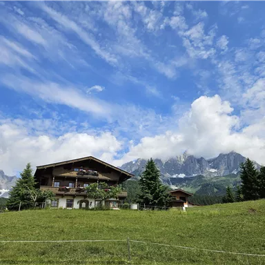 Ein schönes Bauernhaus umgeben von einer grünen Wiese und hohen Bergen. Der Himmel ist klar mit einigen Wolken.