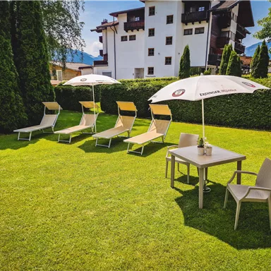 A well-maintained garden with sun loungers and a table under a sun umbrella. In the background, a multi-story building can be seen.