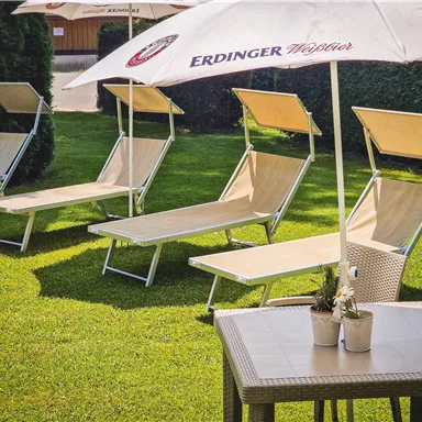 There are several lounge chairs on a green lawn area, equipped with sun umbrellas. Next to the lounge chairs is a table with decorative plants.