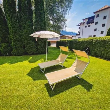 Two loungers and a sunshade are on a green meadow. In the background, tall hedges and a modern building can be seen.