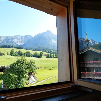 A view from a window onto a green meadow and majestic mountains. In the foreground, a wooden house and trees can be seen.
