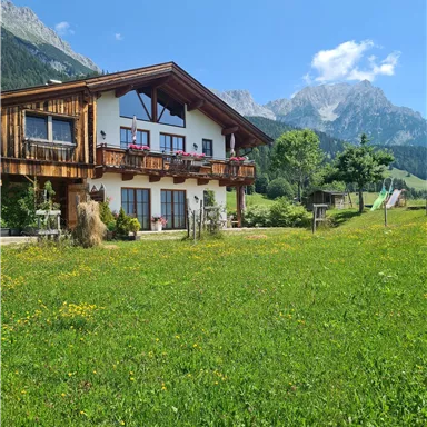 Ein schönes Holzhaus in einer grünen Wiese mit Bergen im Hintergrund. Der Himmel ist klar und blau, was eine idyllische Landschaft schafft.