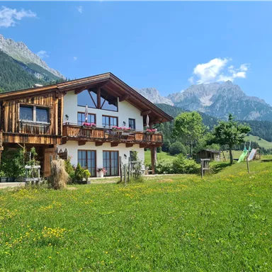 Ein schönes Holzhaus in einer malerischen Berglandschaft. Umgeben von Wiesen und hohen Bergen unter einem klaren blauen Himmel.