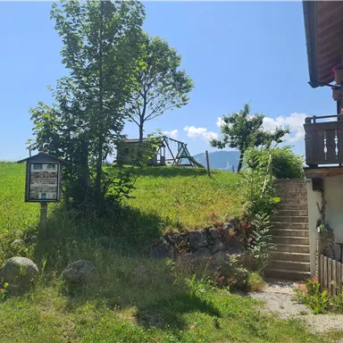 Ein grüner Hang mit einem Spielplatz im Hintergrund und einer Treppe, die zu einem Gebäude führt. Die Sonne scheint an einem klaren Tag.