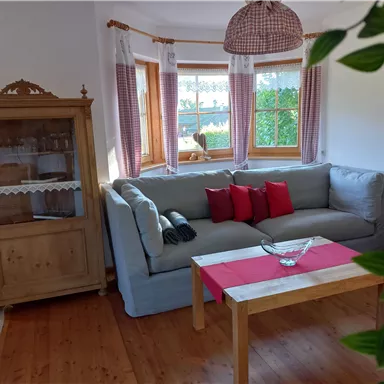 Ein gemütliches Wohnzimmer mit einem bequemen grauen Sofa und roten Kissen. Ein Holztisch steht in der Mitte, umgeben von großen Fenstern, die viel Licht hereinlassen.