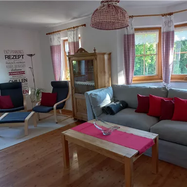 Ein gemütliches Wohnzimmer mit einem hellen Sofa und roten Kissen. Es gibt einen Holztisch und Fenster mit Vorhängen, die viel Tageslicht hereinlassen.