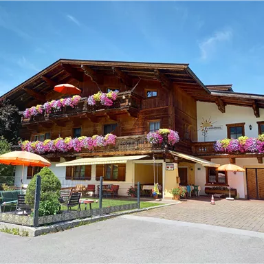 A traditional wooden house with colorful flower balconies and a clear blue sky. In front of the house, there is a garden area and sun umbrellas.