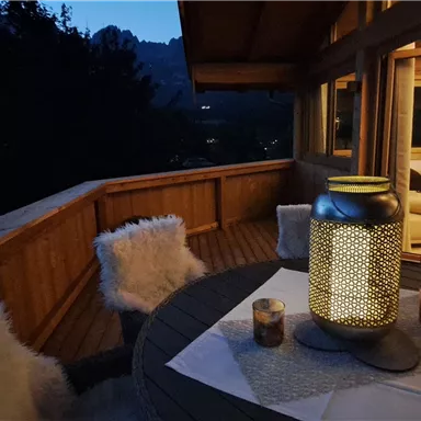 A cozy terrace at night with a table, a lantern, and soft sheep skins. In the background, illuminated windows and mountains can be seen.