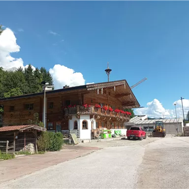 Ein traditionelles Holzhaus mit Balkon und roten Blumen. Im Hintergrund sind Bäume und ein blauer Himmel sichtbar.