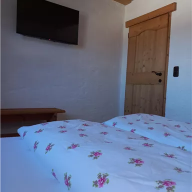 Ein gemütliches Schlafzimmer mit einem Doppelbett und floraler Bettwäsche. An der Wand hängt ein Fernseher, und die Tür aus Holz führt nach draußen.