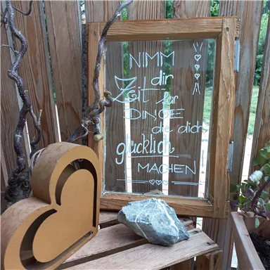 Ein Holzrahmen mit einer handgeschriebenen Botschaft, die dazu auffordert, sich Zeit für die Dinge zu nehmen, die glücklich machen. Daneben liegt eine herzförmige Deko und ein Stein auf einem Holzregal.