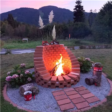Ein gemütlicher Lagerfeuerplatz aus Ziegelsteinen mit brennendem Feuer. Umgeben von Blumen und Gräsern, bietet er eine warme Atmosphäre in der Natur.