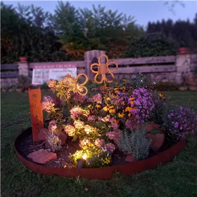 Ein bunter Blumenbeet mit verschiedenen Pflanzen und einer dekorativen Blume aus Holz. Das Beet ist in warmem Licht beleuchtet und befindet sich in einem gepflegten Garten.
