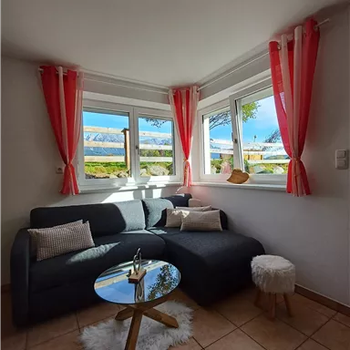 Ein gemütliches Wohnzimmer mit einem grauen Sofa und einem Glastisch. Große Fenster lassen viel Licht herein und bieten einen Blick nach draußen.