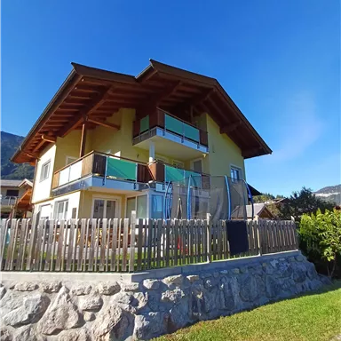 Ein schönes Haus mit Holzveranda und großen Fenstern. Es steht in einer grünen Umgebung unter einem blauen Himmel.