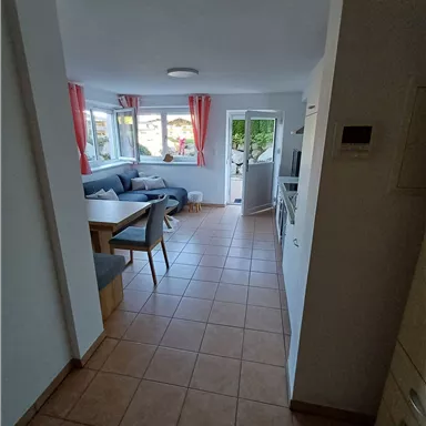 Ein heller Flur mit keramischen Fliesen führt in einen gemütlichen Wohnraum. Der Raum ist modern eingerichtet, mit einem Sofa und großen Fenstern.