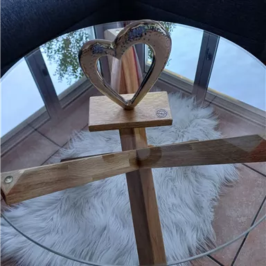 Ein runder Glastisch mit einer Holzbasis in Herzform. Der Tisch steht auf einem weißen Teppich und vor einem Fenster.