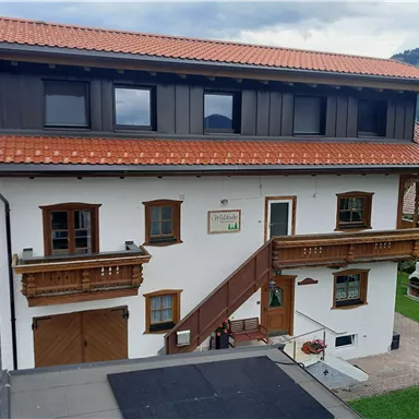 A beautiful, traditional house with a red tiled roof and wooden balconies. It is located in a quiet rural setting with a view of the mountains.