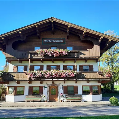 Ein traditionelles alpenländisches Holzhaus mit blühenden Balkonen. Die Umgebung ist grün und sonnig, ideal für einen entspannten Aufenthalt.