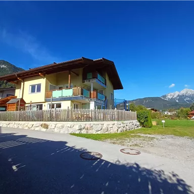 Ein gelbes Haus mit mehreren Balkonen und einem gepflegten Garten. Im Hintergrund sind Berge und ein klarer blauer Himmel zu sehen.