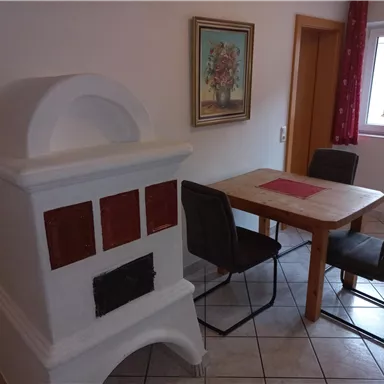 Ein gemütlicher Essbereich mit einem Holzofen und einem Tisch mit Stühlen. An der Wand hängt ein Gemälde, und das Fenster bringt natürliches Licht herein.