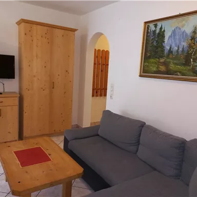 Ein gemütlicher Wohnbereich mit einer grauen Couch, einem Holztisch und einem TV. An der Wand hängt ein Bild von einem Berglandskap.