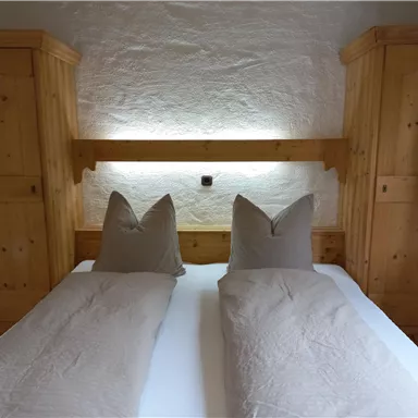 Ein gemütliches Schlafzimmer mit einem Doppelbett und Holzschränken. Die Kissen haben eine besondere Form und es gibt sanftes Licht hinter dem Kopfteil.