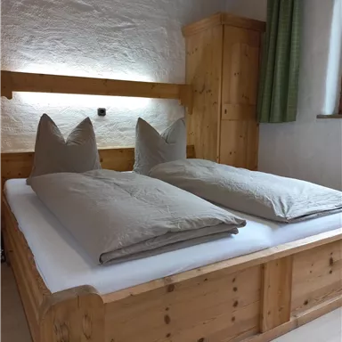 Ein gemütliches Schlafzimmer mit einem Holzbett und großen, grauen Kopfkissen. Die Wände sind hell und es gibt einen Schrank aus Holz sowie einen Vorhang.