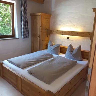 Ein gemütliches Schlafzimmer mit einem Doppelbett aus Holz und grauen Bettdecken. Das Zimmer hat ein Fenster mit Blick ins Grüne und warme Beleuchtung.