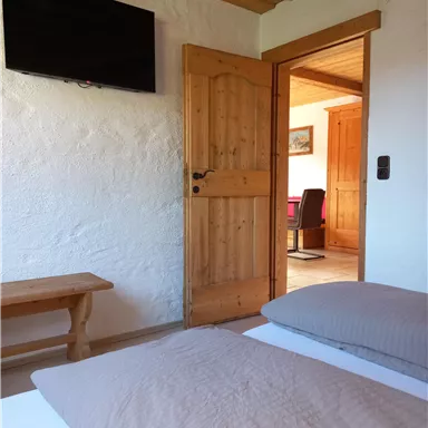 Ein gemütliches Schlafzimmer mit zwei Betten und einem Holzstuhl. Durch die offene Tür sieht man einen weiteren Raum mit einem Schreibtisch.