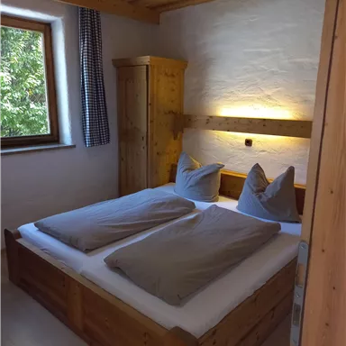 Ein gemütliches Schlafzimmer mit einem Doppelbett aus Holz und großen Kissen. Das Zimmer hat ein Fenster, das Tageslicht hereinlässt.