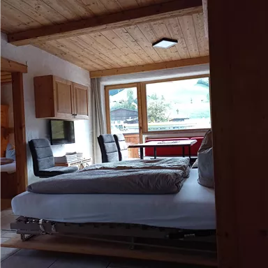 Ein gemütliches Zimmer mit einem Bett und zwei Stühlen. Große Fenster sorgen für viel Licht und Blick auf die Landschaft.