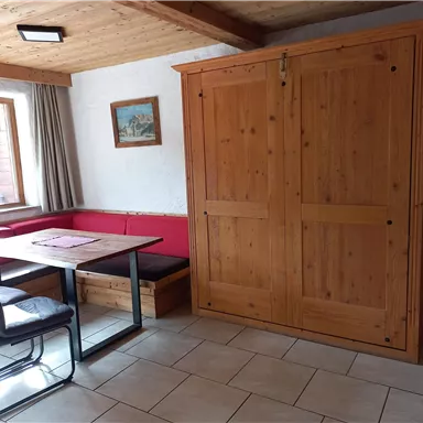 Ein gemütlicher Raum mit einem Tisch und Stühlen. An der Wand steht ein Holzschrank und ein Fenster mit Aussicht.