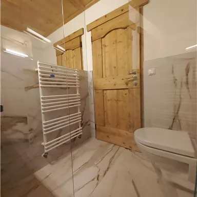 Ein modernes Badezimmer mit marmorierten Fliesen und einer Glasdusche. Zwei Holztüren und ein Handtuchhalter aus Metall sind gut sichtbar.