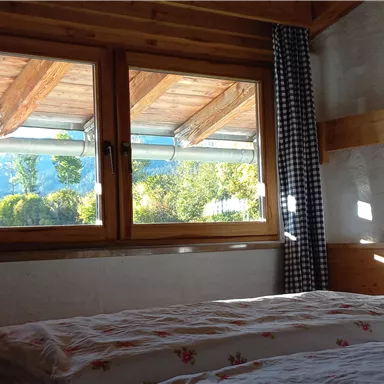 Ein gemütliches Schlafzimmer mit einem Bett und einem Fenster, das einen Blick auf die Natur bietet. Helles Sonnenlicht strömt durch das Fenster und beleuchtet den Raum.