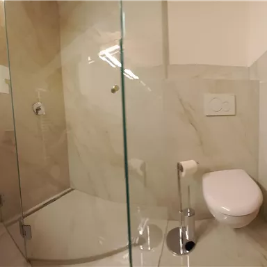 Ein modernes Bad mit einer Duschkabine aus Glas und einer eleganten Toilette. Die Wände sind mit hellen, marmorierten Fliesen verkleidet.