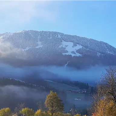 Ein malerischer Berg mit schneebedeckten Gipfeln und Nebel, der über die Hügel zieht. Der Himmel ist klar und blau, was eine ruhige Atmosphäre schafft.