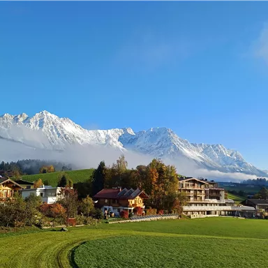 Eine majestätische Berglandschaft mit schneebedeckten Gipfeln und klarem blauen Himmel. Im Vordergrund sind grüne Wiesen und schöne Gebäude zu sehen.