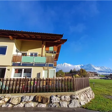 Ein modernes Haus mit Balkon und einer Holzverkleidung. Im Hintergrund sind Berge und ein blauer Himmel zu sehen.
