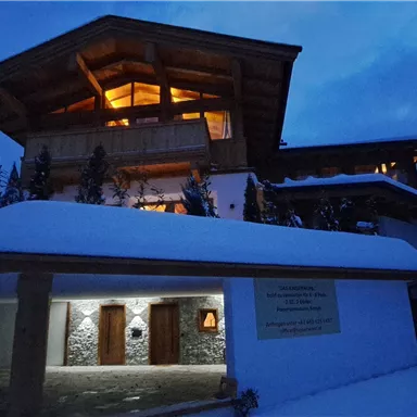 An alpine house at night, surrounded by snow. Warm light streams from the windows, creating an inviting atmosphere.