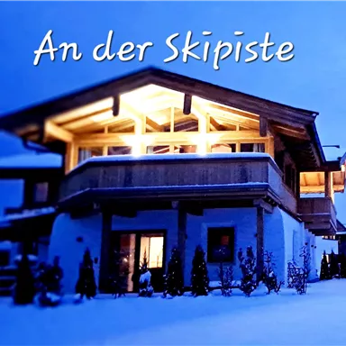 A cozy wooden house in a snow-covered setting. It is evening and the light radiates warmly from the windows.