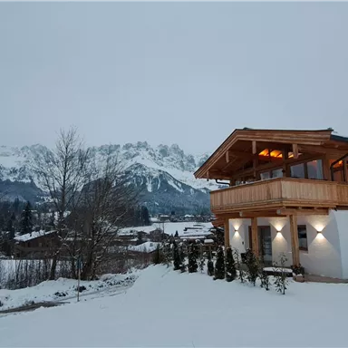A modern house in the snow with a wooden balcony. Snow-covered mountains can be seen in the background.