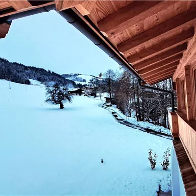A landscape with snow-covered fields and trees. The view is seen from a wooden building, which conveys a cozy atmosphere.